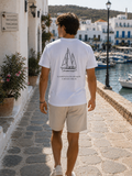 A man walking away on a stone harbor path wearing a white t-shirt with a black sailboat graphic and the text 'A SAILOR'S NAVY: A MILED MAST.' The background shows a sunny coastal town with white buildings, boats in the water, and green hills.