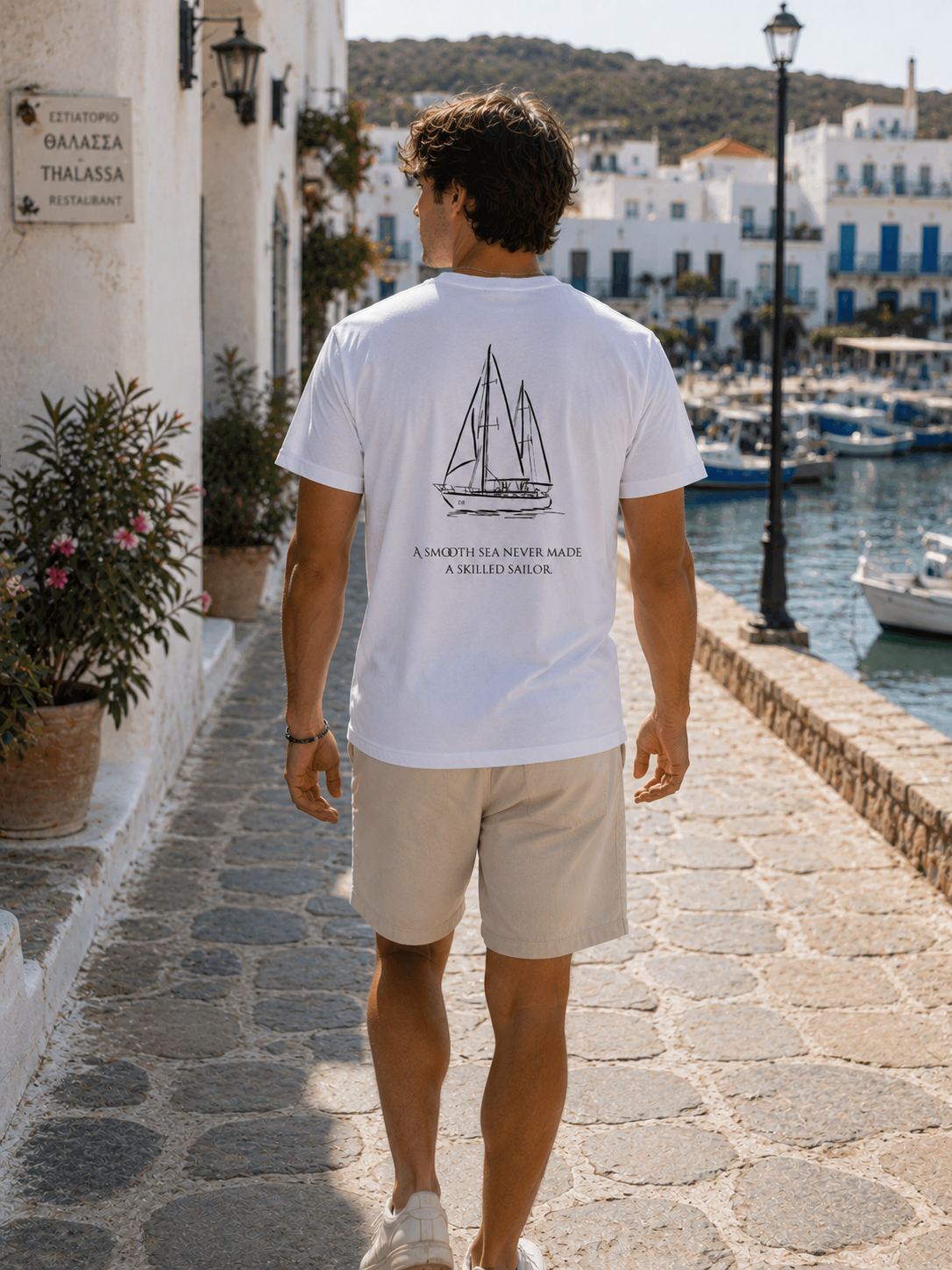A man walking away on a stone harbor path wearing a white t-shirt with a black sailboat graphic and the text 'A SAILOR'S NAVY: A MILED MAST.' The background shows a sunny coastal town with white buildings, boats in the water, and green hills.